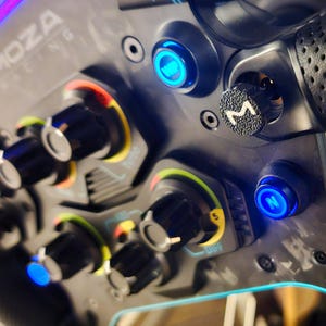 May include: Close-up of a black racing wheel with multiple buttons and knobs. The wheel has a blue and white logo that says "MOZA RACING".