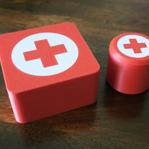 May include: Two red first aid boxes with a white cross on the top. The larger box is square and the smaller box is round.