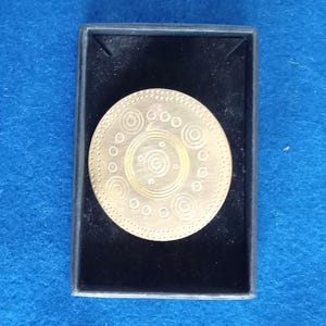 May include: A gold-colored metal brooch with a circular design featuring concentric circles and dots. The brooch is displayed in a black velvet box.