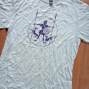 May include: A light sage green short-sleeved t-shirt featuring a dark purple circular graphic on the chest. The design depicts a central figure holding two long spears and a round shield, flanked by two small dog-like creatures within the circle.