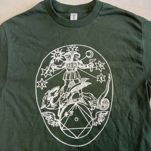 May include: A dark green t-shirt featuring a white graphic print of a two-headed figure riding a dragon. The figure is holding a staff and a sword. The graphic is enclosed within a circle containing stars and a crescent moon.