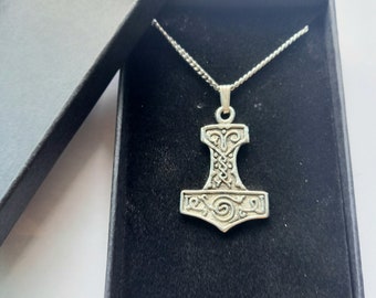 Op de afbeelding: Een zilverkleurige hangerketting met Thor's hamer, Mjolnir, gepresenteerd in een zwarte doos. De hamerhanger heeft ingewikkelde Keltische knoopwerk en een spiraalvormig ontwerp. De ketting is ook zilver.