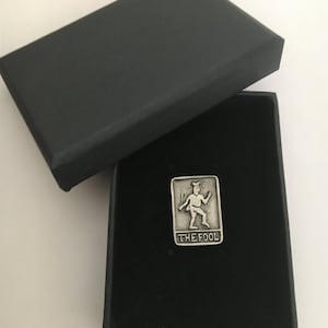 May include: A silver pewter tarot card charm featuring The Fool card from the Major Arcana. The charm is in a black velvet box.
