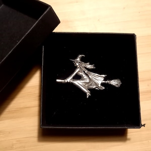 May include: A silver-toned witch figurine riding a broomstick, displayed in a black velvet box.