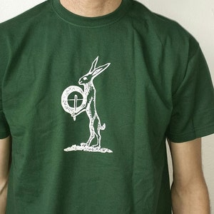 Puede incluir: Una camiseta verde oscuro con un gráfico blanco de un conejo sosteniendo un círculo con una cruz dentro. El conejo está de pie sobre sus patas traseras y tiene orejas largas.