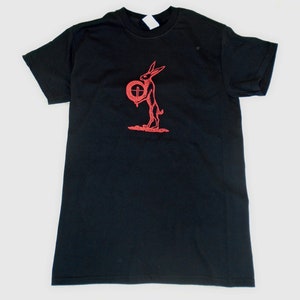 Puede incluir: Camiseta negra con un gráfico rojo de un conejo sosteniendo un objeto circular con una cruz. El conejo está de pie. La camiseta está hecha de un material suave.