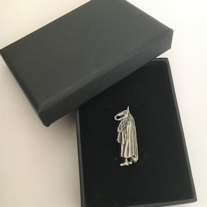 Op de afbeelding: Een zilverkleurige speld met een figuur met een paardenkop en een gedrapeerde mantel, gepresenteerd in een open zwarte doos met een zwart interieur. De speld is gedetailleerd en valt op tegen de donkere achtergrond.