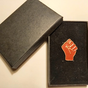 Può includere: Uno spillo in smalto rosso con un pugno chiuso in una scatola di velluto nero. Lo spillo è un simbolo di solidarietà e resistenza.