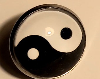 May include: A silver-toned pin with a black and white yin yang symbol. The yin yang symbol is a circular design with a black half and a white half, each containing a smaller circle of the opposite color.