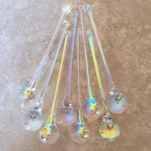 May include: A collection of seven clear glass icicle ornaments, each featuring a spherical base and a long, tapered stem. The glass has an iridescent, rainbow-like finish that reflects light, creating a shimmering effect across the surface.
