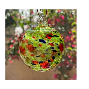 May include: A round, clear glass ornament with a green base color and colorful flecks of red, blue, and orange. The ornament is suspended by a thin, gold-colored wire. The background is blurred, showing pink flowers.