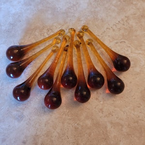 May include: A collection of ten amber-colored glass teardrop ornaments. Each ornament has a rounded bulbous base that tapers into a slender stem, topped with a small loop. The glass has a gradient effect, transitioning from a deep brown to a lighter amber.