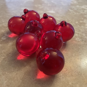 May include: A collection of seven small, translucent red glass spheres, each featuring a tiny, integrated loop at the top. The smooth, polished surfaces of these round ornaments reflect light, creating bright highlights across their deep crimson color.