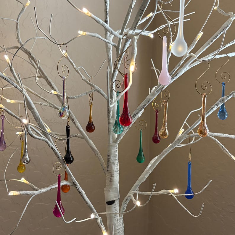 May include: A collection of teardrop-shaped glass ornaments in various colors including red, blue, green, yellow, pink, and clear, each hanging from a gold-toned metal spiral hook on a white artificial tree with integrated warm white LED lights.