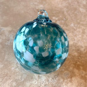 May include: A clear, teal-colored glass ornament with white, irregular shapes. The ornament has a loop at the top for hanging. The surface has a textured, mosaic-like appearance. This decorative item is suitable for holiday or home decor.