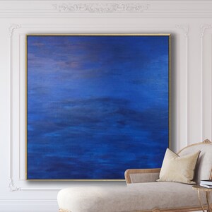 May include: Large square abstract painting with shades of blue and gold, framed in a gold border. The artwork hangs on a white wall, with a chaise lounge and side table in the foreground. The painting's colors create a calming and serene atmosphere.