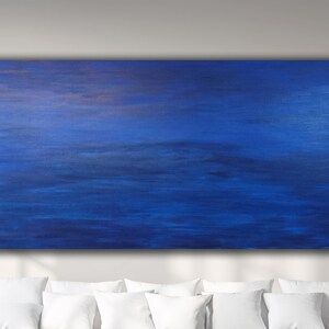 May include: A large, rectangular abstract painting with a predominantly blue color palette. The artwork features horizontal brushstrokes in varying shades of blue, creating a sense of depth and movement. The painting is mounted on a wall above white pillows.