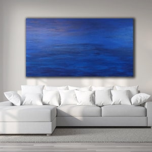 May include: Large abstract painting with shades of blue and a hint of gold. The artwork hangs above a white sectional sofa with white pillows. A lamp and side table are to the right.