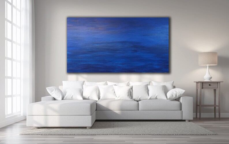 May include: Large abstract painting with shades of blue and a hint of gold. The artwork hangs above a white sectional sofa with white pillows. A lamp and side table are to the right.