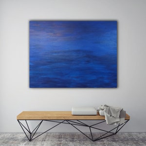 May include: An abstract painting in shades of blue hangs above a wooden bench with a black metal geometric base. The painting has a textured appearance, with darker blue tones at the top and lighter shades below. A white cushion and a gray blanket rest on the bench.