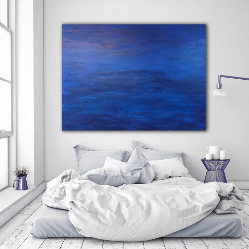 May include: A large, rectangular abstract painting in shades of blue hangs above a bed with white bedding. The painting has a textured appearance, with darker blue tones at the bottom and lighter hues at the top. The room is bright and minimalist.