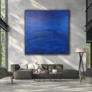 May include: A large, square abstract painting dominates a modern living room. The artwork features a gradient of deep blues, creating a sense of depth and movement. The room includes a gray sectional sofa, two armchairs, and a floor lamp, all set against a white brick wall.