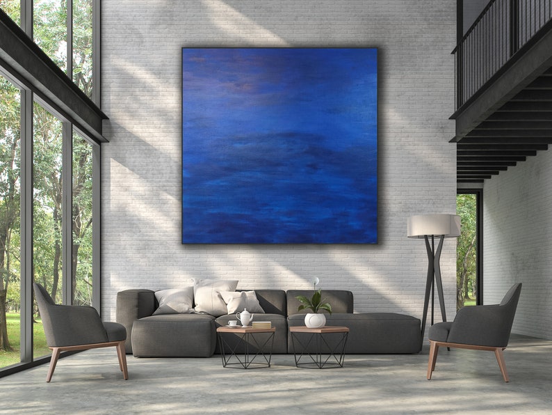 May include: A large, square abstract painting dominates a modern living room. The artwork features a gradient of deep blues, creating a sense of depth and movement. The room includes a gray sectional sofa, two armchairs, and a floor lamp, all set against a white brick wall.