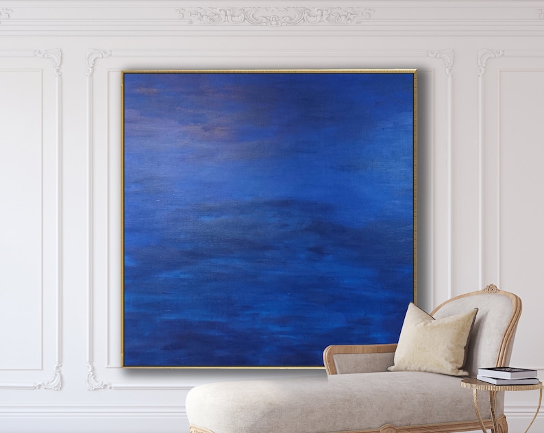 May include: Large square abstract painting with shades of blue and gold, framed in a gold border. The artwork hangs on a white wall, with a chaise lounge and side table in the foreground. The painting's colors create a calming and serene atmosphere.