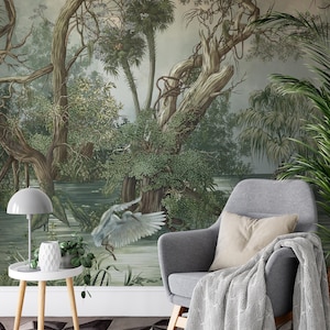 Op de afbeelding: Een serene interieurscène met een gedetailleerde muurschildering van een moeraslandschap met bomen, water en een vogel in vlucht. Een grijze fauteuil met een deken, een bijzettafel met een lamp en vaas, en een geometrisch patroon tapijt.