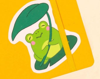 Könnte beinhalten: Ein grüner Frosch-Aufkleber mit geschlossenem Auge, der ein grünes Blatt über seinem Kopf hält. Der Aufkleber befindet sich auf einem gelben Notizbuch mit einem gelben Gummiband.