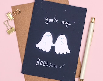 Op de afbeelding: Een marineblauwe wenskaart met de tekst "You're my" en "BOOOOOOO..." met twee witte spookillustraties die elkaars hand vasthouden. Een gouden pen en paperclips zijn ook aanwezig.
