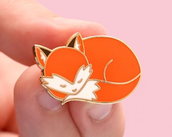 May include: A gold-colored enamel pin featuring a cartoon fox curled up asleep. The fox is orange with white accents on its face and tail.
