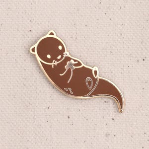 Op de afbeelding: Een bruine emaille pin van een otter die een klein bloemetje vasthoudt. De otter is wit omlijnd en heeft een gouden afwerking.
