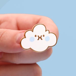 May include: A gold-coloured enamel pin shaped like a white cloud with a cute cartoon face. The cloud has two blue eyes and a small frown.
