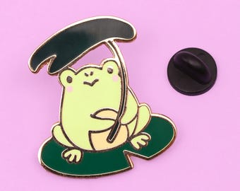 May include: A green frog with a black umbrella sits on a green lily pad. The frog is holding the umbrella with its left hand. The frog has a yellow belly and a pink blush on its cheeks.