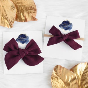 May include: Two burgundy fabric hair bows with a white card behind them. The card has the text "stitched arrow" in gold lettering on a blue watercolor background.