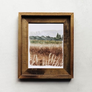 May include: A framed watercolor landscape print. The artwork depicts a field of tall, dry grass in shades of brown and gold, with a backdrop of rolling hills and trees. The frame is made of rustic, weathered wood.