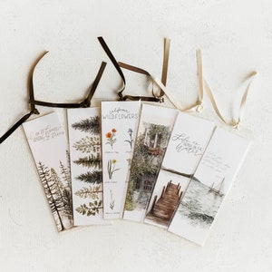 Könnte beinhalten: Eine Sammlung von sechs rechteckigen Lesezeichen mit Illustrationen zum Thema Natur und Text. Die Lesezeichen zeigen Bilder von Bäumen, Wildblumen, einer Szene am Wasser und Texten wie "I'm at home in the trees". Jedes Lesezeichen hat ein Band.