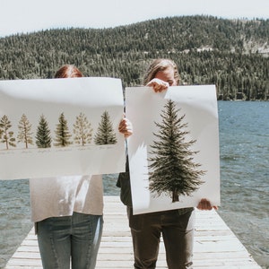 May include: Two people are holding up large prints of trees. The print on the left shows a row of seven different types of trees. The print on the right shows a single large tree.