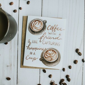 Könnte beinhalten: Eine weiße Karte mit brauner Schrift, auf der steht "Coffee with a friend is like capturing happiness in a cup". Die Karte zeigt zwei Aquarell-Illustrationen von Kaffeetassen mit Latte Art.
