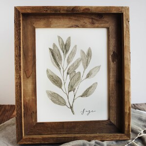 May include: A framed watercolour painting of a sage plant with green leaves. The frame is made of rustic wood and has a white mount. The word "Sage" is written in cursive at the bottom of the painting.