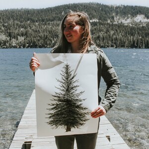 May include: A person holding a white poster with a black and green illustration of a pine tree. The poster is being held in front of a lake and a forest.