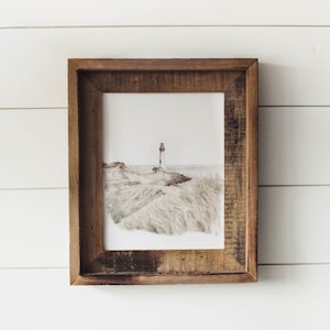 May include: A framed print of a lighthouse on a grassy hill overlooking the ocean. The print is in a rustic wooden frame.