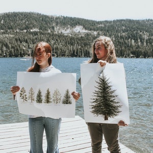 May include: Two people stand on a wooden dock holding up posters of trees. The poster on the left shows a row of five different types of trees with their names written below. The poster on the right shows a single large tree.