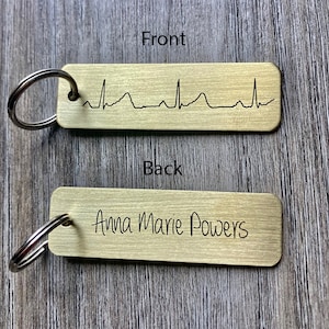Puede incluir: Llavero de metal dorado con una línea de ritmo cardíaco en la parte delantera y el nombre "Anna Marie Powers" grabado en la parte posterior.