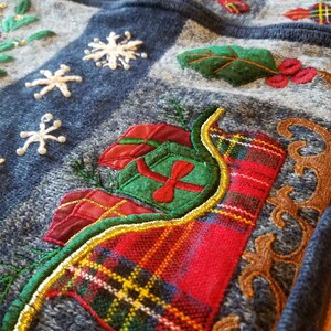 May include: A blue knitted fabric with embroidered Christmas designs. The design features a red and green plaid sleigh with a green and red gift box, a gold trim, and a brown ornate design.