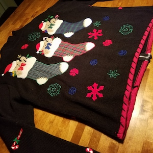 May include: Black Christmas sweater with a red, green, and white candy cane design on the sleeves. The sweater features three embroidered bears wearing stocking caps and holding candy canes, inside of stockings with plaid patterns. The sweater is decorated with snowflakes and other festive designs.