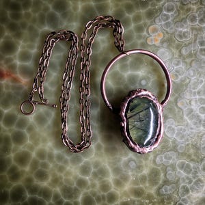 May include: A handcrafted necklace featuring a large, oval green gemstone pendant with dark inclusions, set in a copper-colored metal frame. The pendant hangs from a copper-toned chain with a toggle clasp. The necklace is displayed on a green, textured surface.