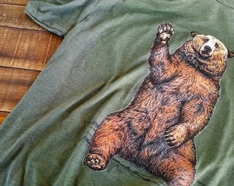 Könnte beinhalten: Ein grünes T-Shirt mit einem braunen Bären-Motiv. Der Bär winkt mit seiner rechten Pfote.