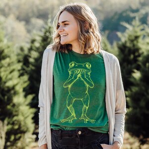 May include: A woman wearing a green t-shirt with a yellow outline of a frog covering its eyes with its hands. The woman is smiling and wearing a white cardigan.